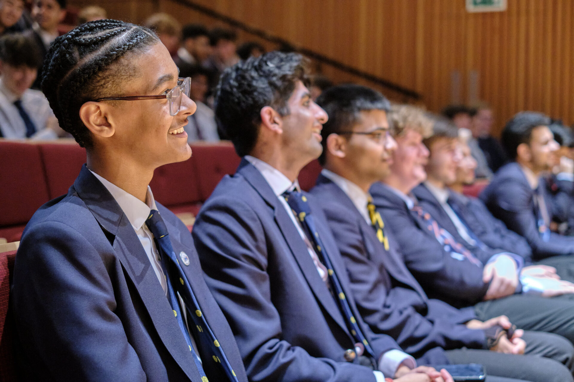 A group of young people in navy blue blazers and ties are seated in a tiered auditorium, looking towards the front.