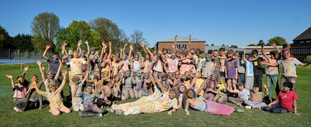 A large group of people covered in colourful powder are posing for a photo on a grassy field under a clear blue sky. Many of them have their arms raised in celebration of Holi.