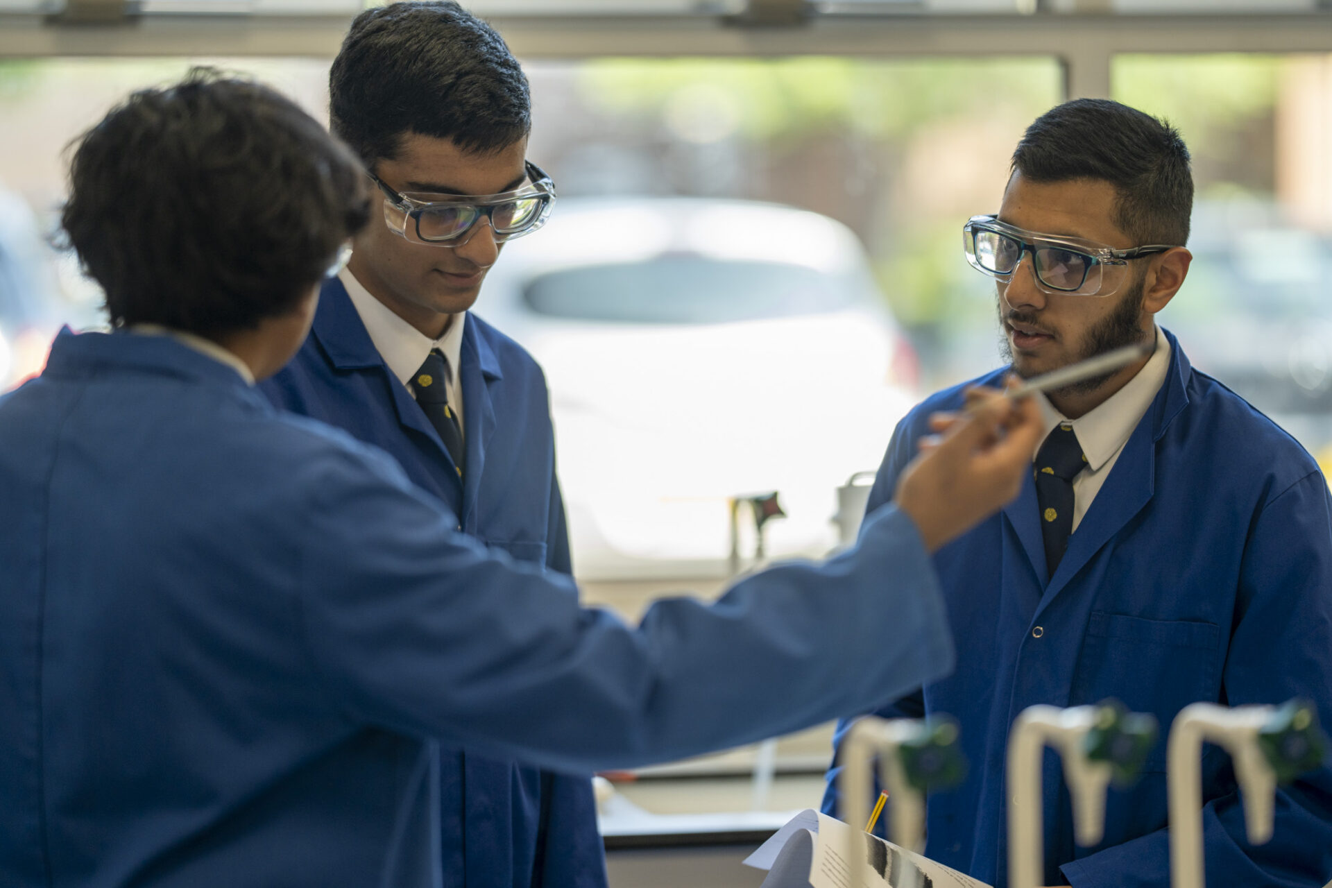 Three students in blue lab coats and safety goggles are gathered around a lab bench, looking at a test tube.