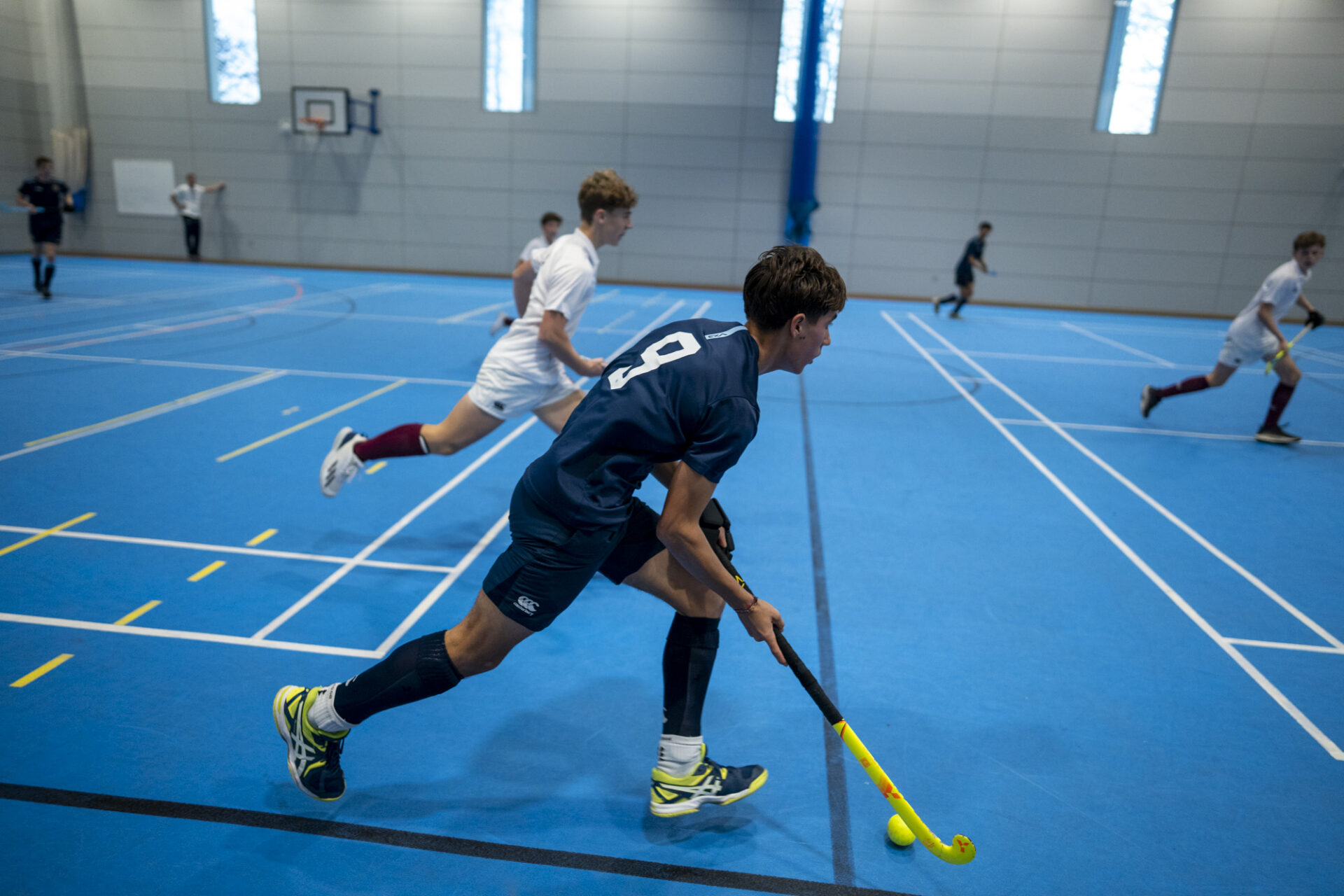 A hockey player chasing a ball across a bright blue indoor court while other players are in the background.