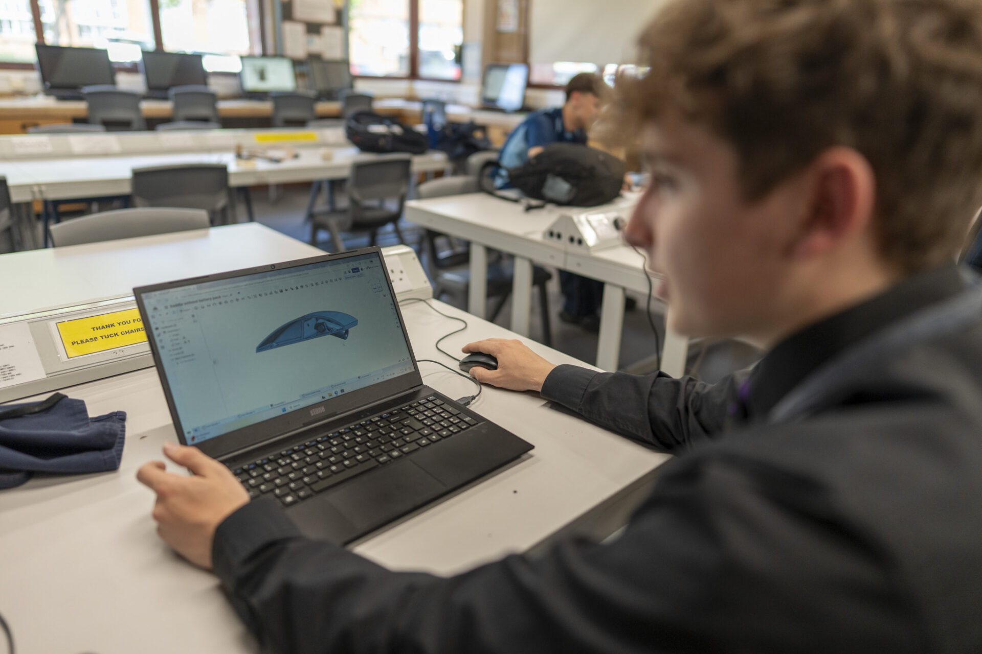 A pupil is using a laptop to view a 3D model of a blue object in a classroom.