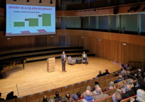 A man stands on a stage in front of a large screen displaying a bar chart about the global sports economy, while another man sits in an armchair to his right. An audience is seated in the foreground.