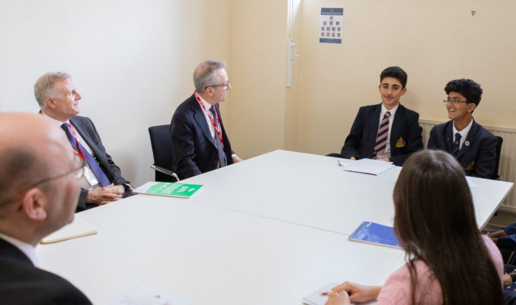 A group of people sitting around a conference table, including two adults in suits and two pupils smiling