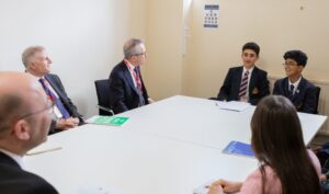 A group of people sitting around a conference table, including two adults in suits and two pupils smiling
