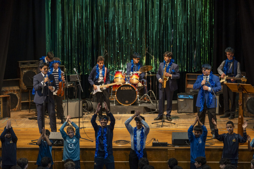 A band of young musicians performs on a stage with a shimmering green backdrop, while a group of boys in the foreground clap along.