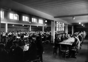 A large group of boys, are seated at long tables in a dining hall. The scene is from a historical period.