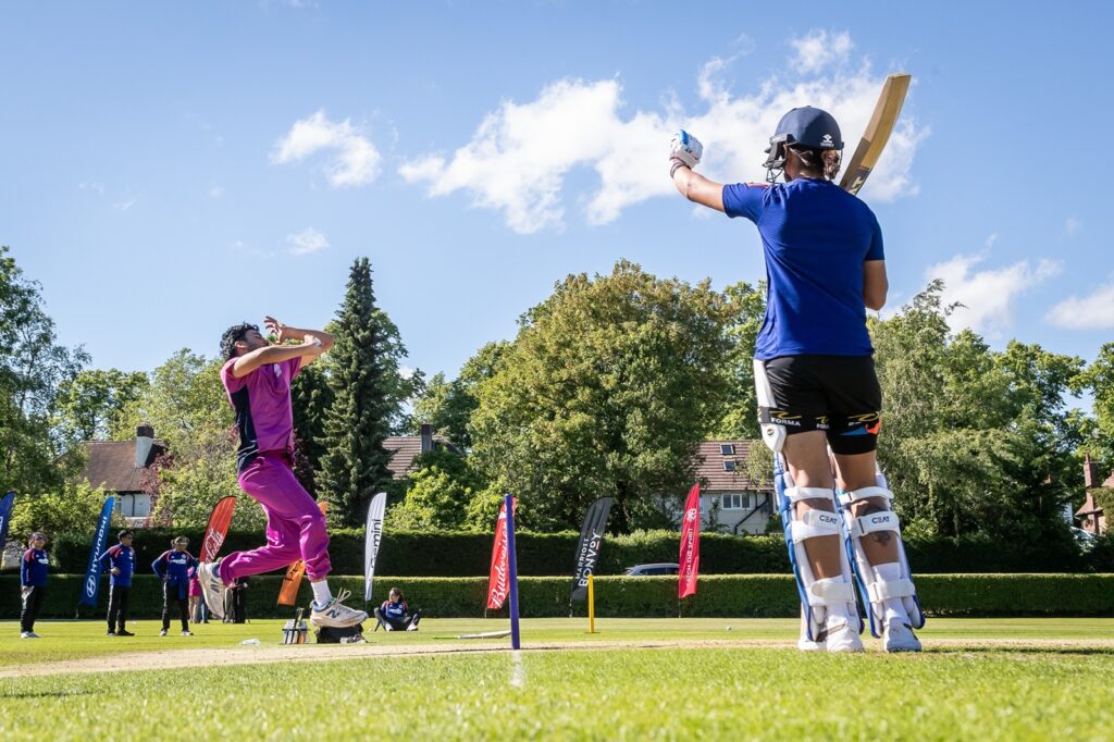 Student bowler in his bowling action watched on by India player holding her bat aloft