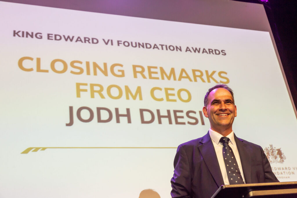 A man in a suit smiles at a podium in front of a screen that reads "KING EDWARD VI FOUNDATION AWARDS CLOSING REMARKS FROM CEO JODH DHESI".