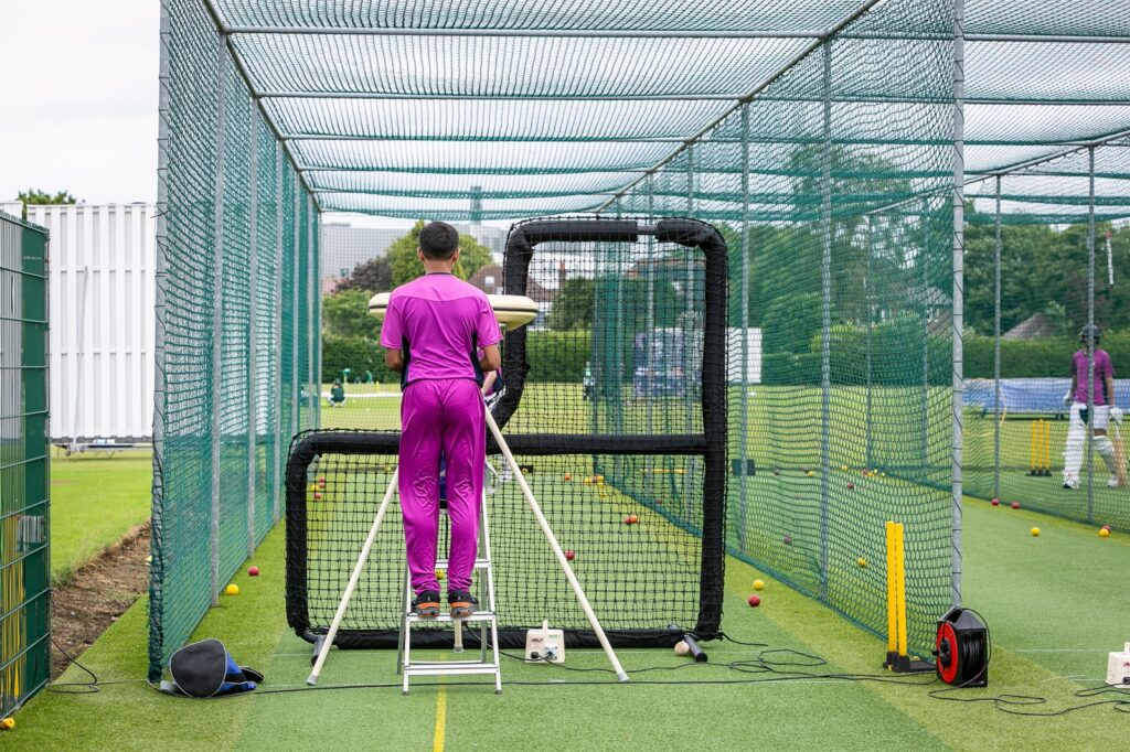 Student in purple net bowlers kit standing on a step ladder by a bowling machine
