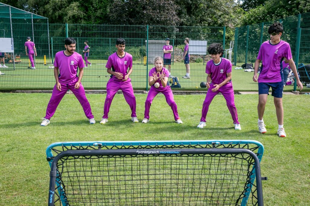 Three male and one female student cricket in purple kit catching a ball thrown against a net