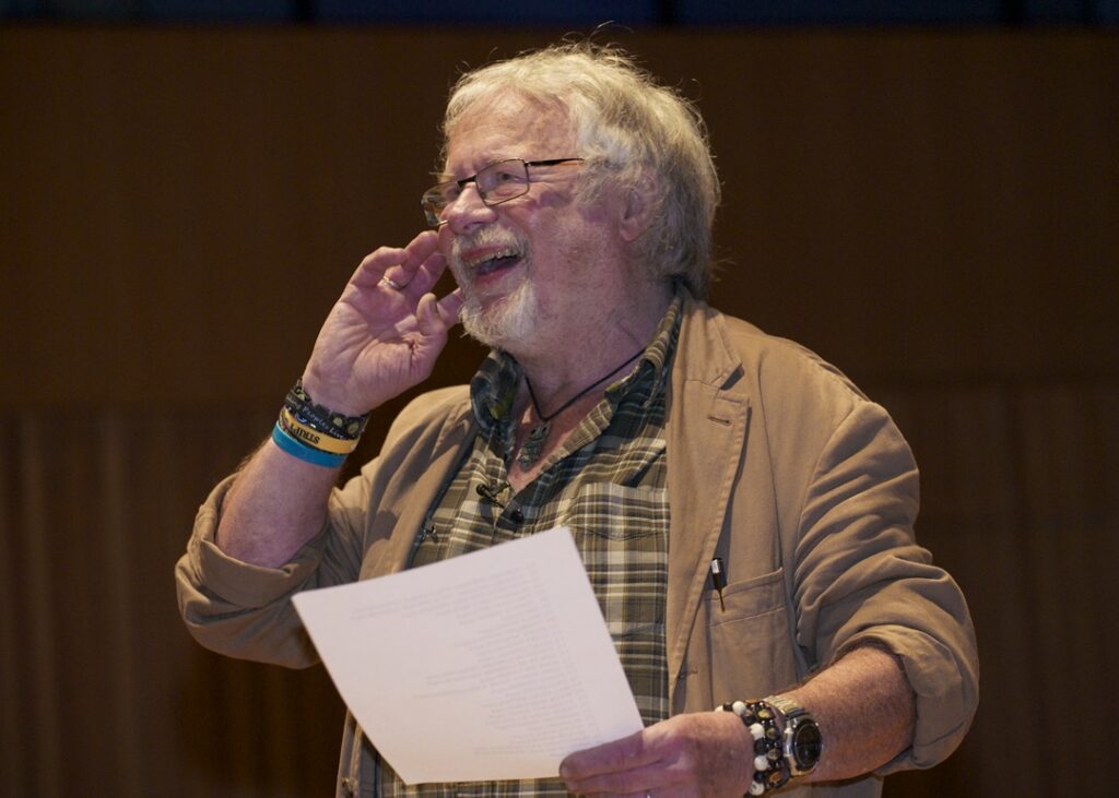 Broadcaster, comedian, writer and conservationist Bill Oddie speaking at KES in 2015. Wearing a light brown jacket and khaki/cream checked shirt, smiling his hand raised to the side of his face.