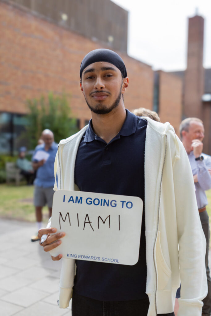 A boy holding a sign that says "I AM GOING TO MIAMI" and "KING EDWARD'S SCHOOL".