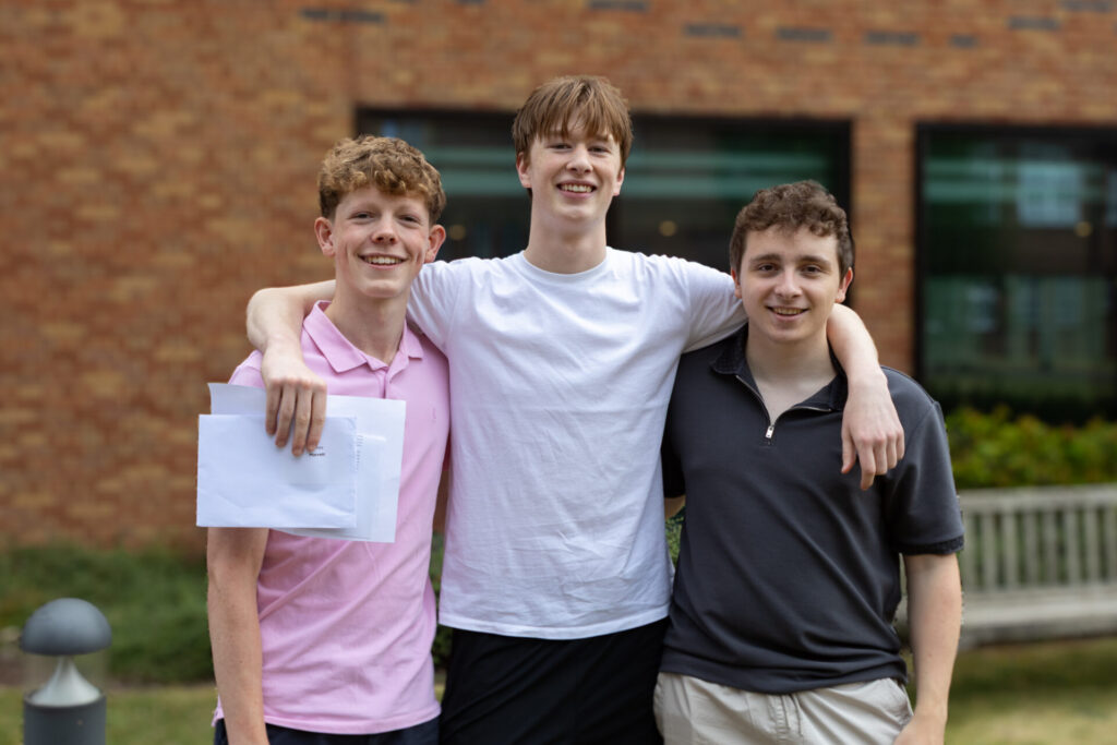 Three smiling individuals stand together outside a brick building, with one holding several envelopes.