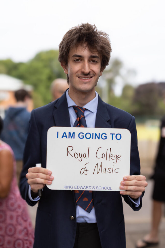 A person in a suit and tie holds a sign that reads "I AM GOING TO Royal College of Music" and "KING EDWARD'S SCHOOL".