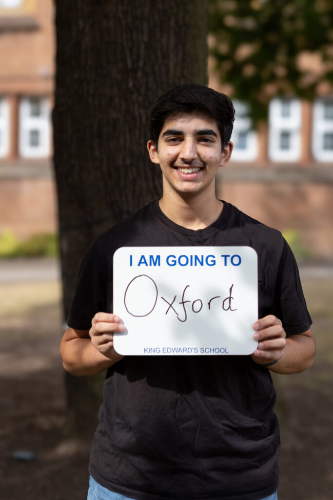 A boy smiles while holding a sign that reads "I AM GOING TO Oxford King Edward's School".