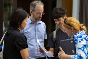 Four people, two adults and two young adults, are gathered around looking at papers and a tablet device.