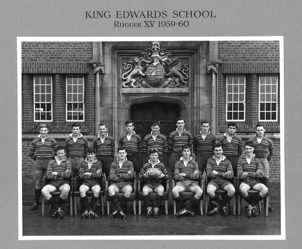 Black and white photograph from 1959/60, showing the KES rugby team. Two rows of pupils - one standing and one sitting. Pupils wearing rugby jerseys and shorts. Bill Oddie seated in the front row holding a rugby ball.