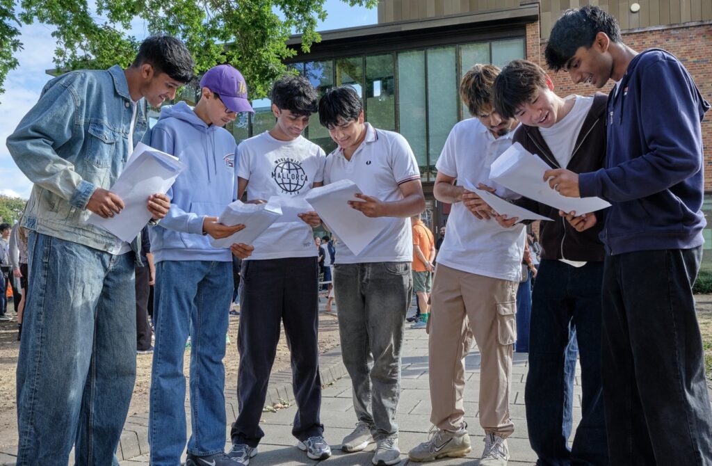 A group of students stand outdoors in a sunny courtyard, reading and holding exam results.