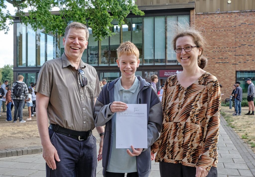 Three people stand outside a campus building, smiling as one students holds a their exam results.