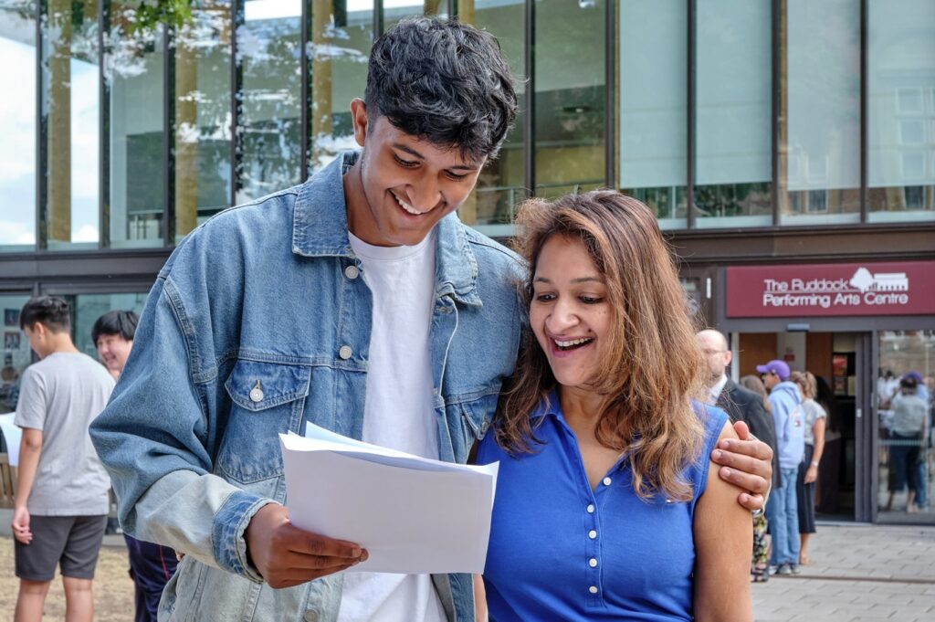 Two people outside while one points at and holds their exam results.