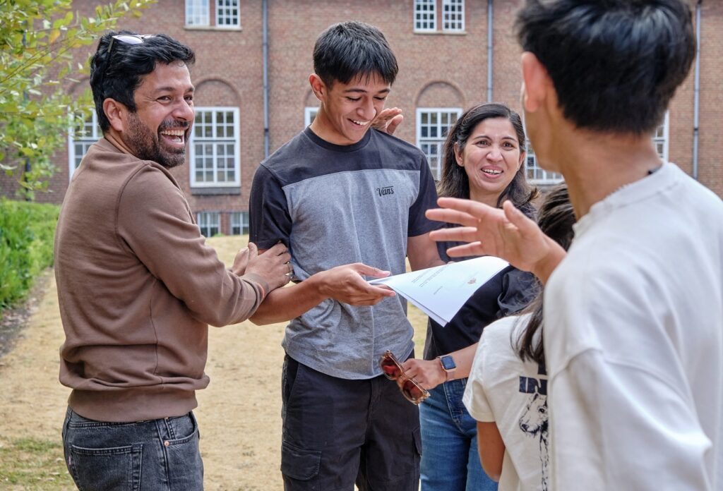 A group of people chatting outdoors smiling while one student holds his exam results.