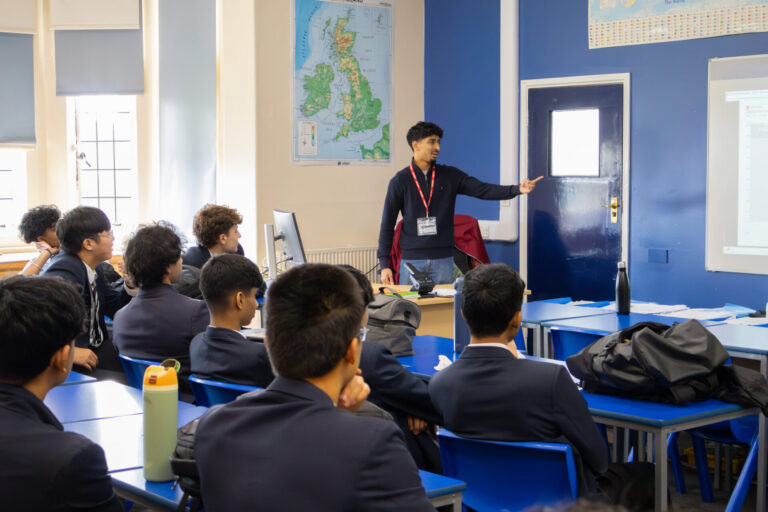 A presenter stands at the front of a classroom, gesturing toward a door while students desks.