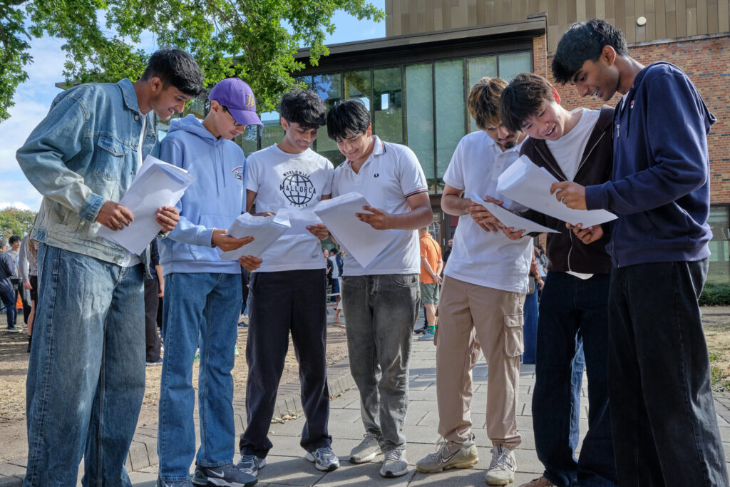 A group of pupils standing outside on a sunny day, each reading their results from a sheet of paper.