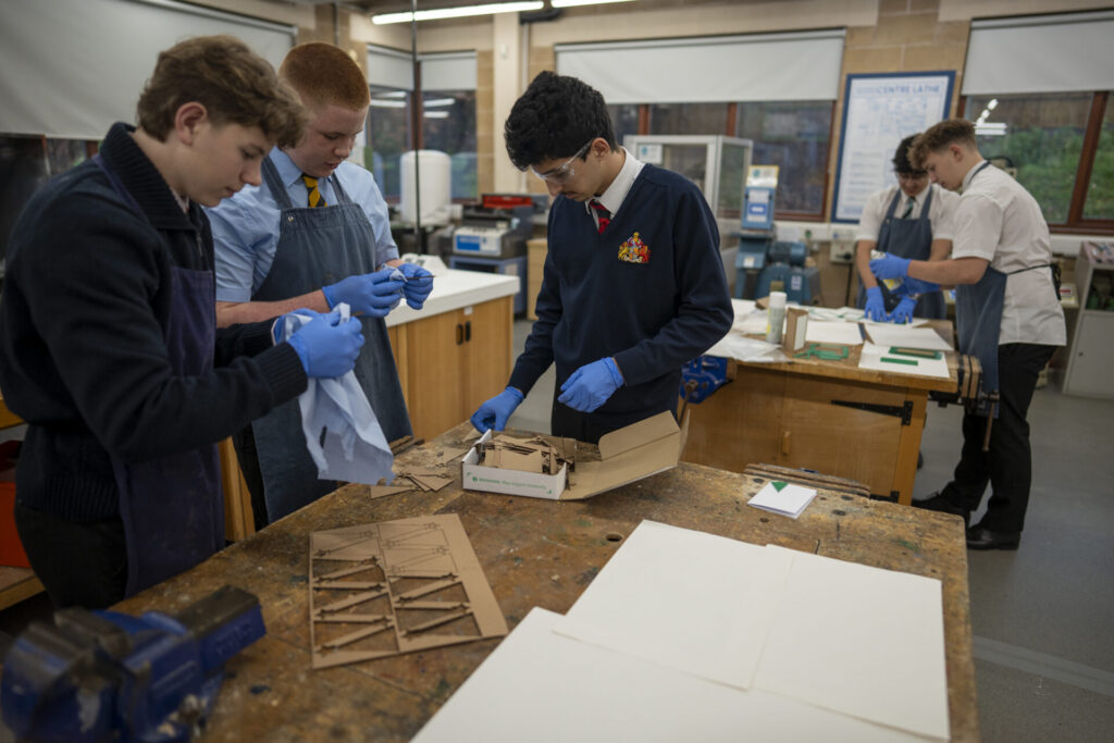 Pupils in a Design and Technology classroom wear blue gloves and work at wooden lab tables preparing materials with tools.