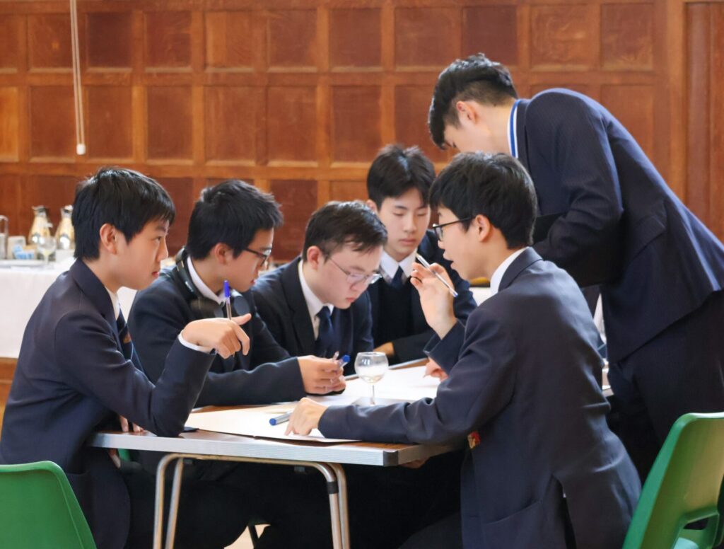 A group of students gather around a table, leaning in and pointing as they discuss a task during an activity in the Enterprise Exchange.