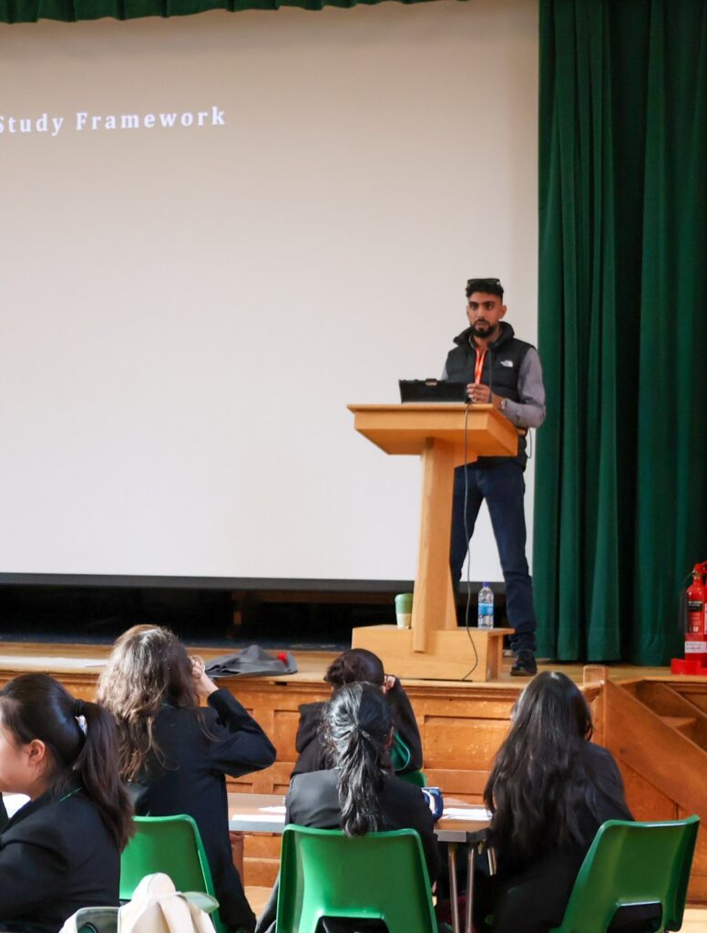 A speaker stands in hall, addressing an audience seated in, with a projection screen behind him and the words “Study Framework” visible.