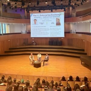 Three speakers sit on stage facing an audience; a large banner above reads “The KE Enterprise Exchange Session 1: Breakfast Briefing.”