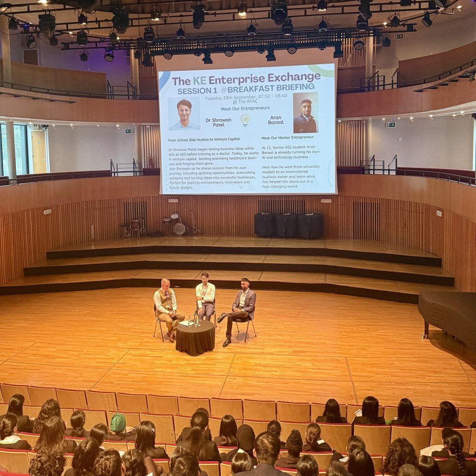 Three speakers sit on stage facing an audience; a large banner above reads “The KE Enterprise Exchange Session 1: Breakfast Briefing.”
